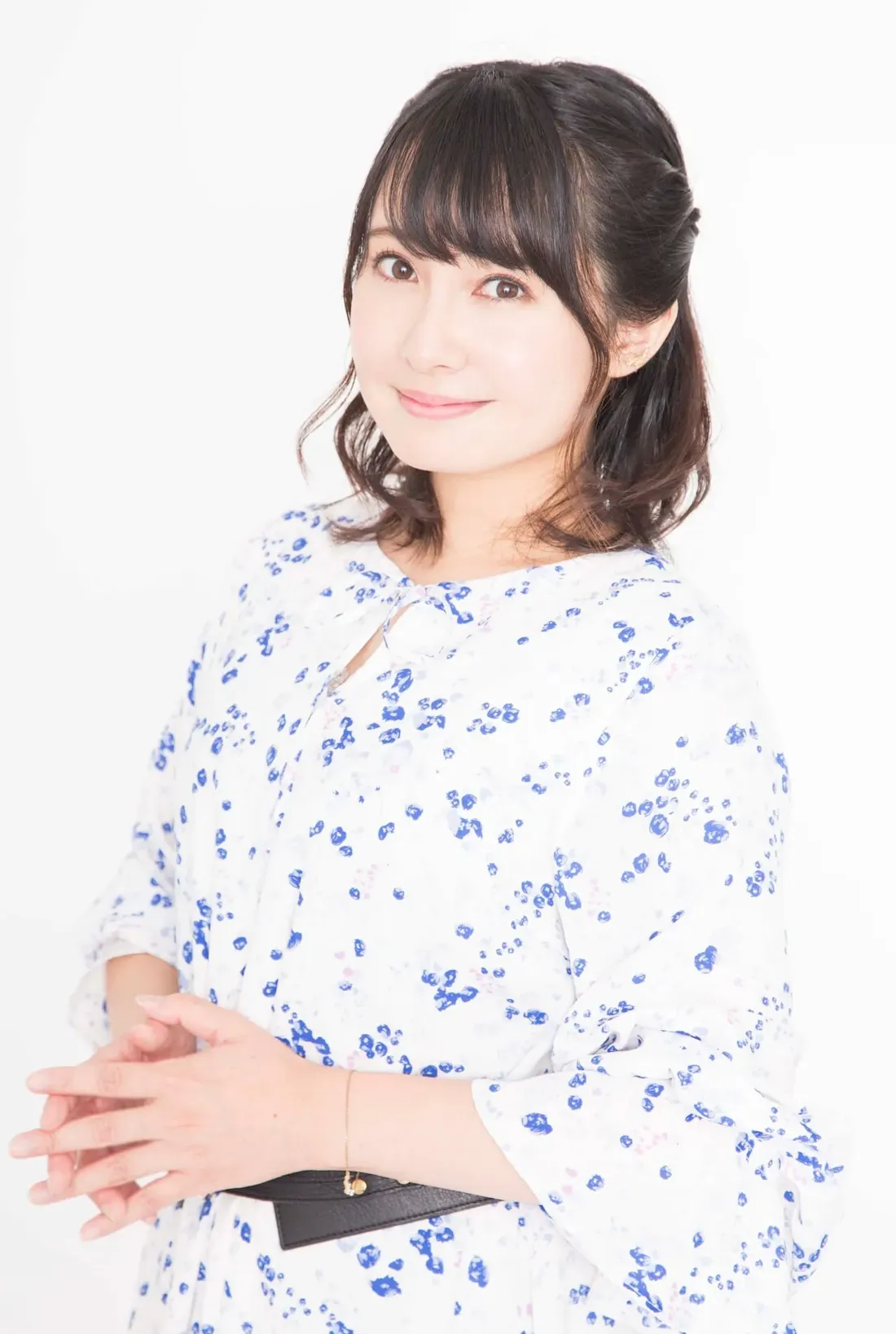 A medium shot of a young East Asian woman with shoulder-length dark brown hair styled with side-swept bangs. She's wearing a white dress adorned with a scattered blue floral pattern.  The dress has three-quarter sleeves with a slightly flared cuff, and a simple keyhole neckline. A thin black belt cinches her waist. Her hands are clasped gently in front of her, and she wears a delicate gold bracelet on her left wrist. She's looking directly at the camera with a pleasant, soft smile. The background is plain white, creating a simple and clean portrait-style image. The overall style is clean, bright, and reminiscent of contemporary portrait photography.
