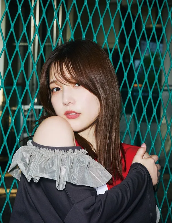 Close-up portrait of a young woman with shoulder-length brown hair, styled with a fringe, posing against a teal diamond-patterned wire mesh fence. She's wearing a black, off-the-shoulder top with a delicate, light grey ruffled trim at the neckline. Underneath, a hint of red and white is visible. Her arms are folded across her body, gently holding onto her upper arms, and she looks directly at the camera with a soft, slightly serious expression. Her makeup is natural and understated. The background is blurred but shows the continuation of the wire fence, suggesting an outdoor setting, possibly an urban or industrial area. The image style is contemporary and fashion-focused, with a slightly moody atmosphere.