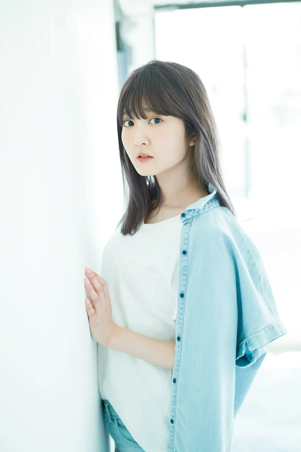 A portrait-style photograph of a young woman with shoulder-length dark brown hair styled with bangs, standing slightly angled to the left, her gaze directed towards the camera.  She is wearing a white, short-sleeved t-shirt and a light blue denim shirt draped over her shoulders.  Her hand rests gently against a bright white wall. The background is blurred and suggests a bright, possibly indoor, setting with a hint of light coming from a window or doorway. The overall style is clean, bright, and reminiscent of a fashion or promotional photograph.