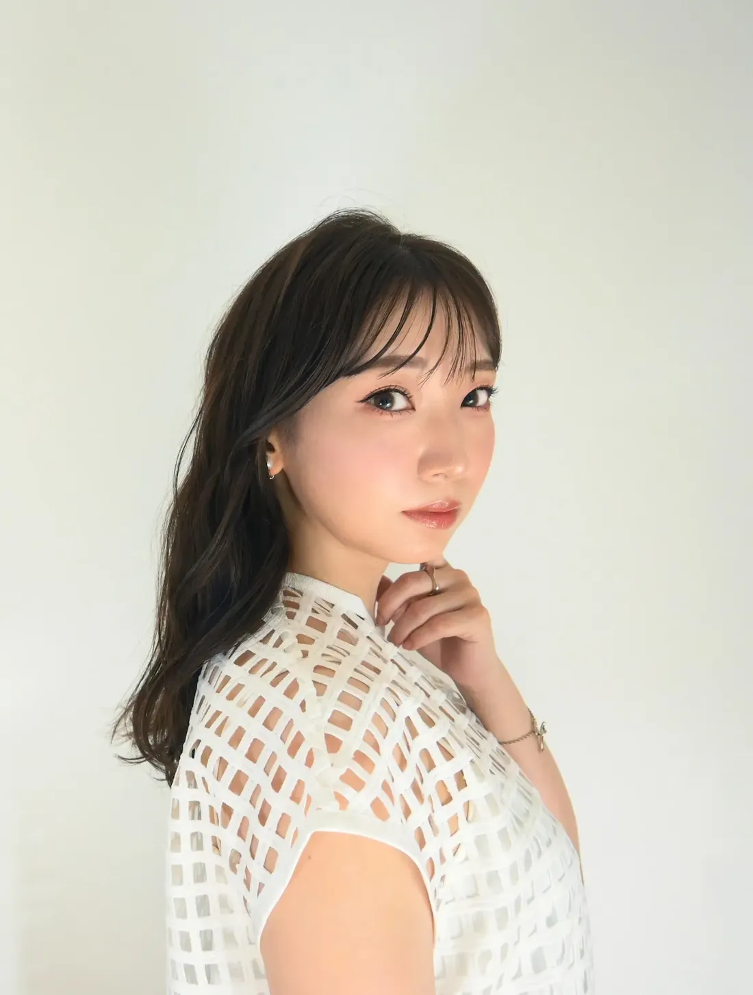 Close-up portrait of a young woman with shoulder-length dark brown hair styled with bangs, glancing over her shoulder. She's wearing a white short-sleeved top with a geometric cut-out pattern, and a delicate silver bracelet on her left wrist. Her hand rests gently on her chin. The background is a plain, off-white wall. The image style is a clean, bright portrait photograph, possibly for a fashion or beauty context.